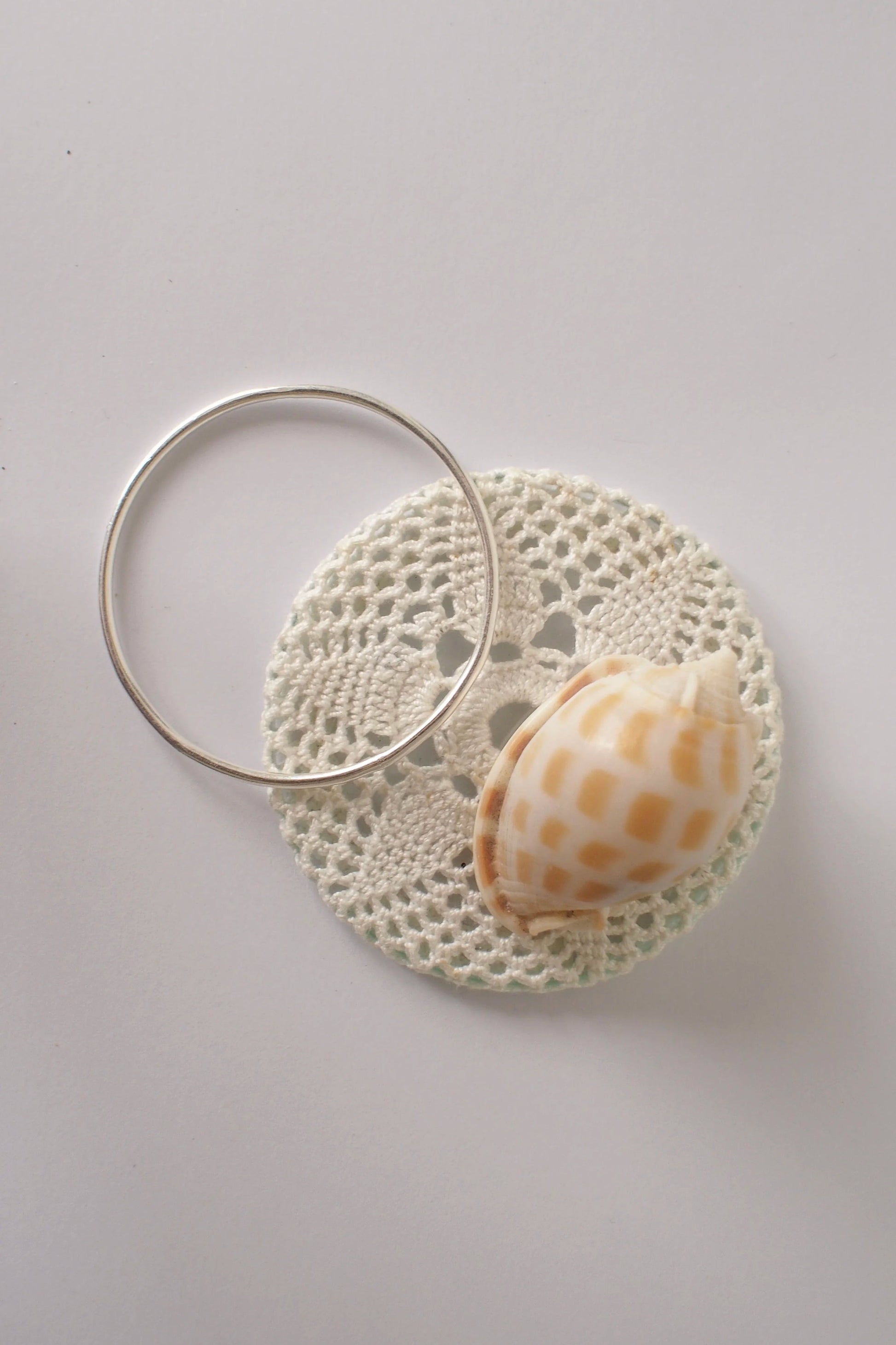 Shell on a doily with a silver bangle on a light gray background