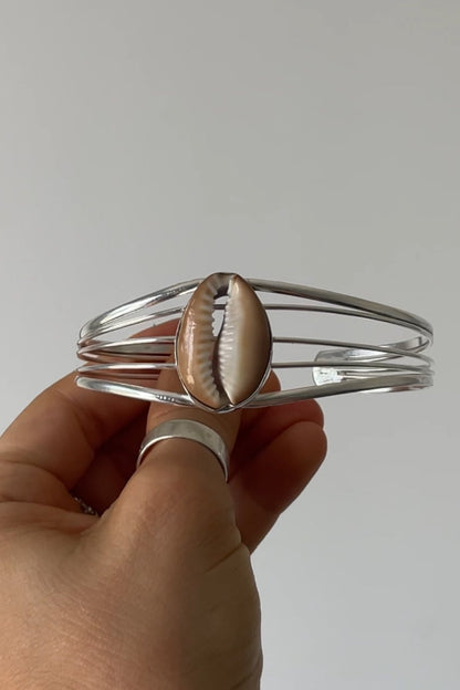 Hand holding a silver bangle with a shell charm against a plain background
